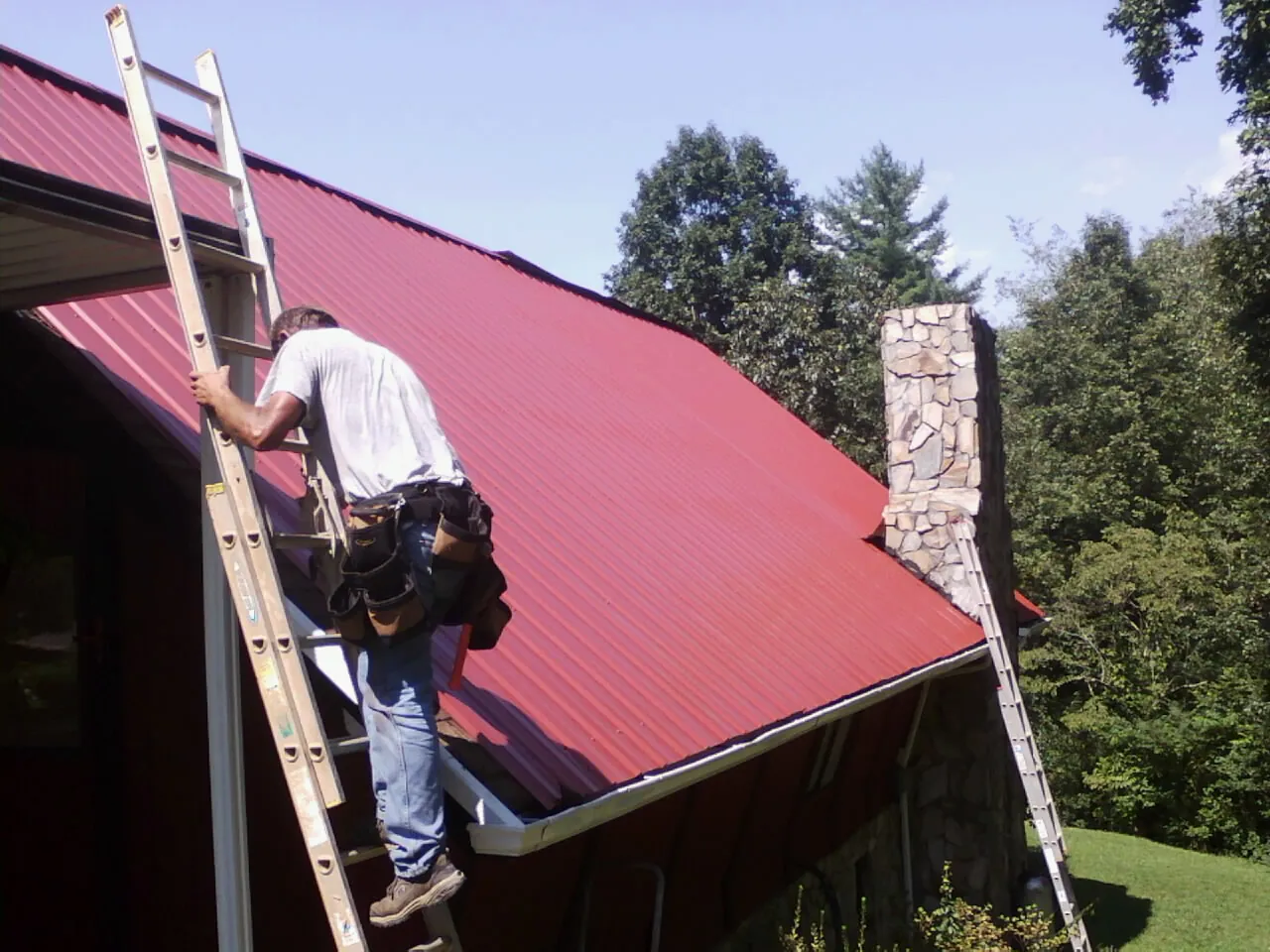 Roofer on a ladder beside a red metal roof and stone chimney — Soffit & Fascia Repair by Roofing Brothers in New Bavaria